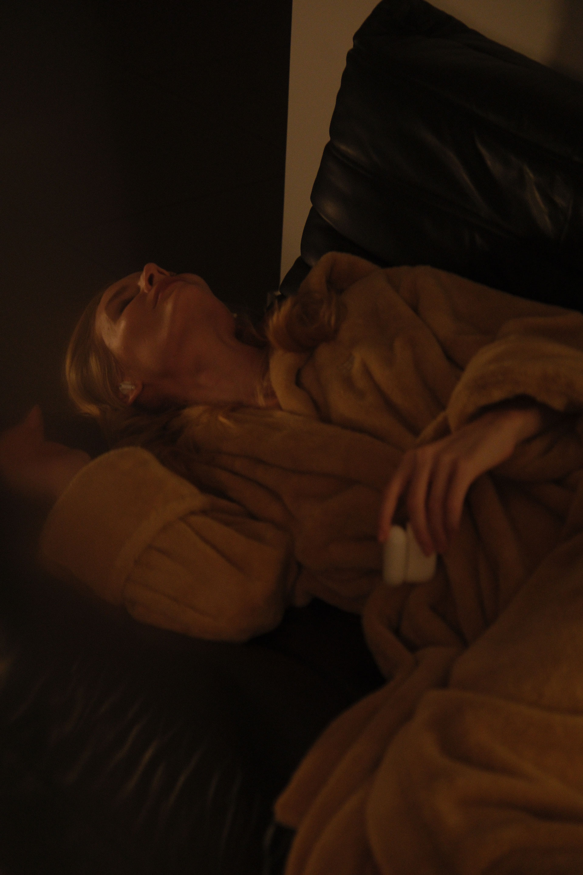 Woman reclining in a dark leather chair wearing a soft beige robe, eyes closed in a calm, restful moment under warm ambient lighting.
