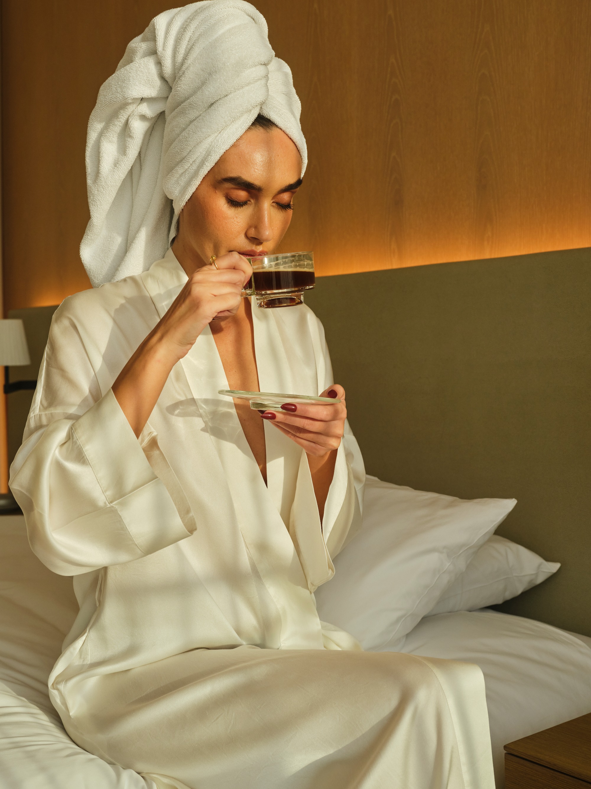 A woman sits on a luxury hotel bed in a silk dressing gown sipping coffee in a glass cup. the next-level wellness platform WELLTTH